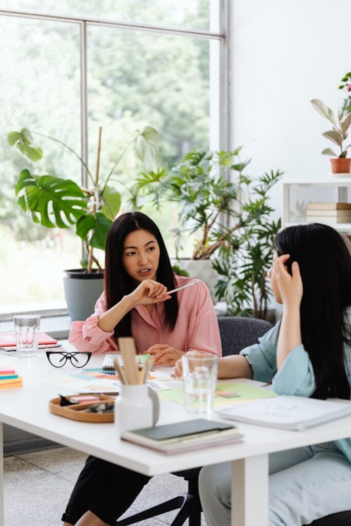 Two Asian women discussing work at an office table with plants around.